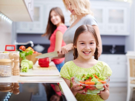 Portrait of happy girl with vegetable salad on background of her mother and sister cooking