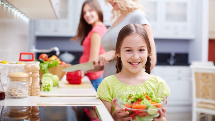 Portrait of happy girl with vegetable salad on background of her mother and sister cooking